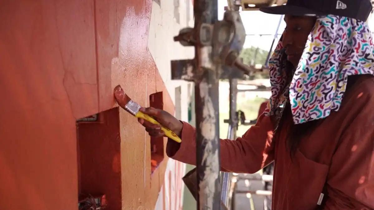Person painting a wall with a red brush, wearing a colorful headscarf and brown jacket.
