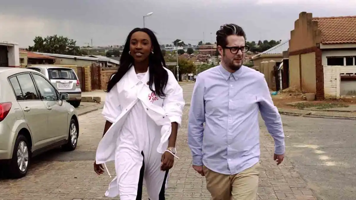 Two people walking down a residential street, with houses and parked cars in the background.