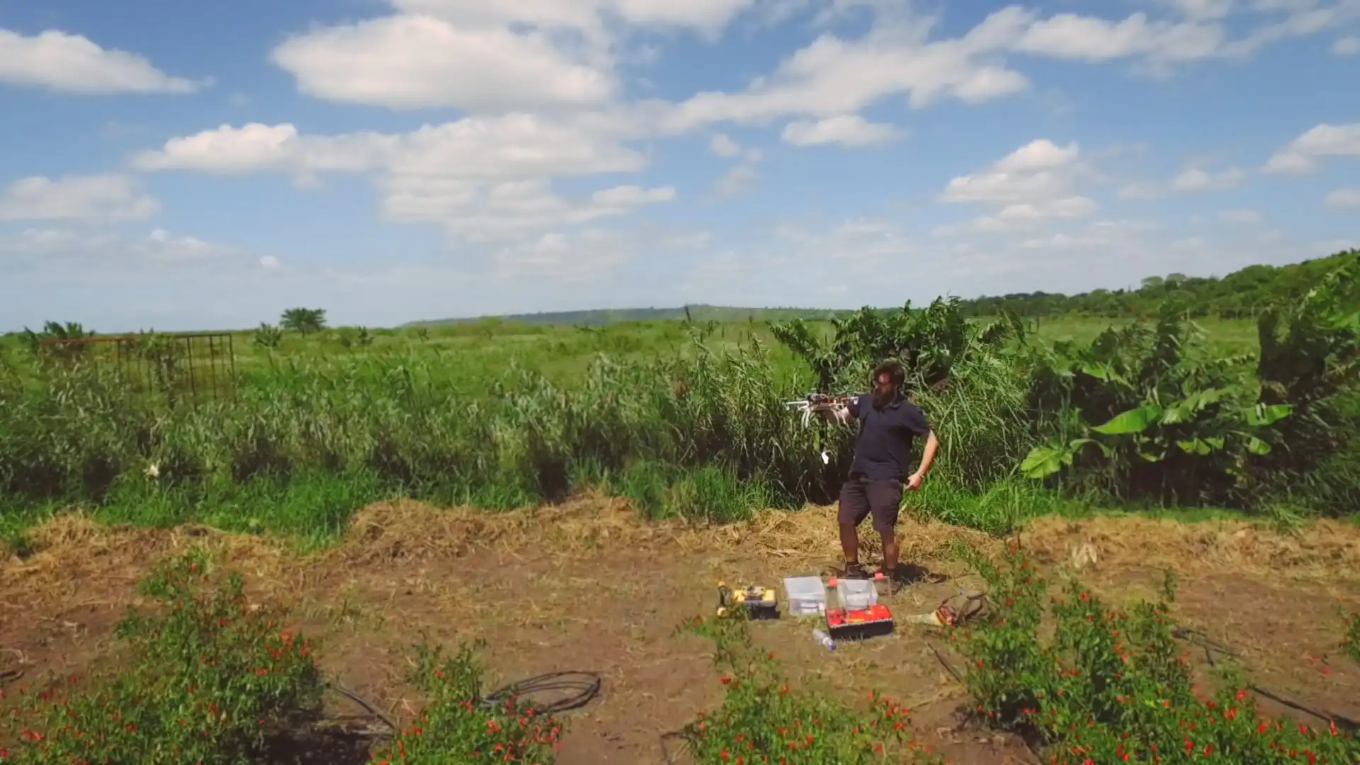 Person operating a drone in a lush field with scattered equipment and greenery under a partly cloudy sky.