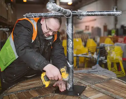 Worker in a warehouse assembling a table using a drill, surrounded by stacked chairs and wrapped furniture.