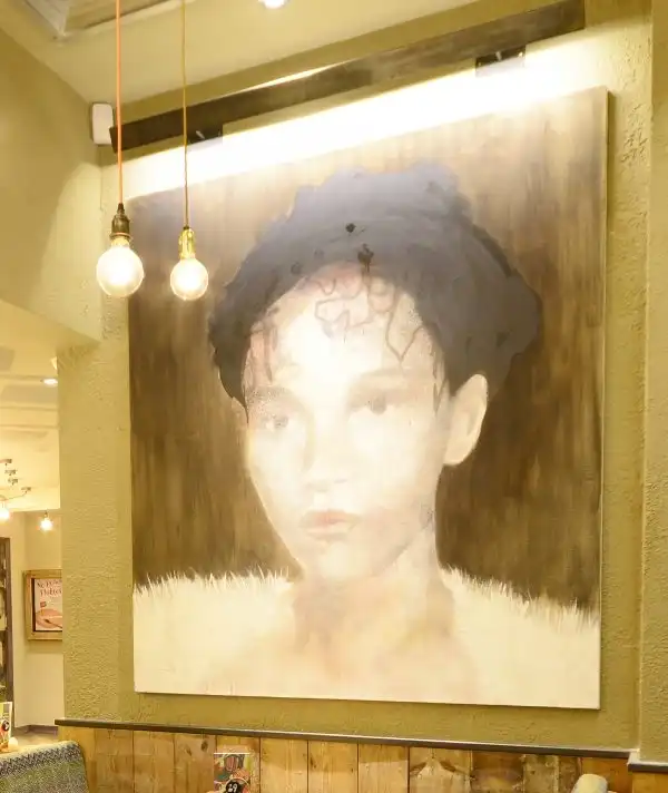 A large portrait painting of a person with short hair, displayed in a warmly lit room with hanging light bulbs.