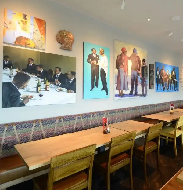 Restaurant interior with vibrant paintings on the wall, wooden tables, and chairs. Bright, artistic atmosphere.